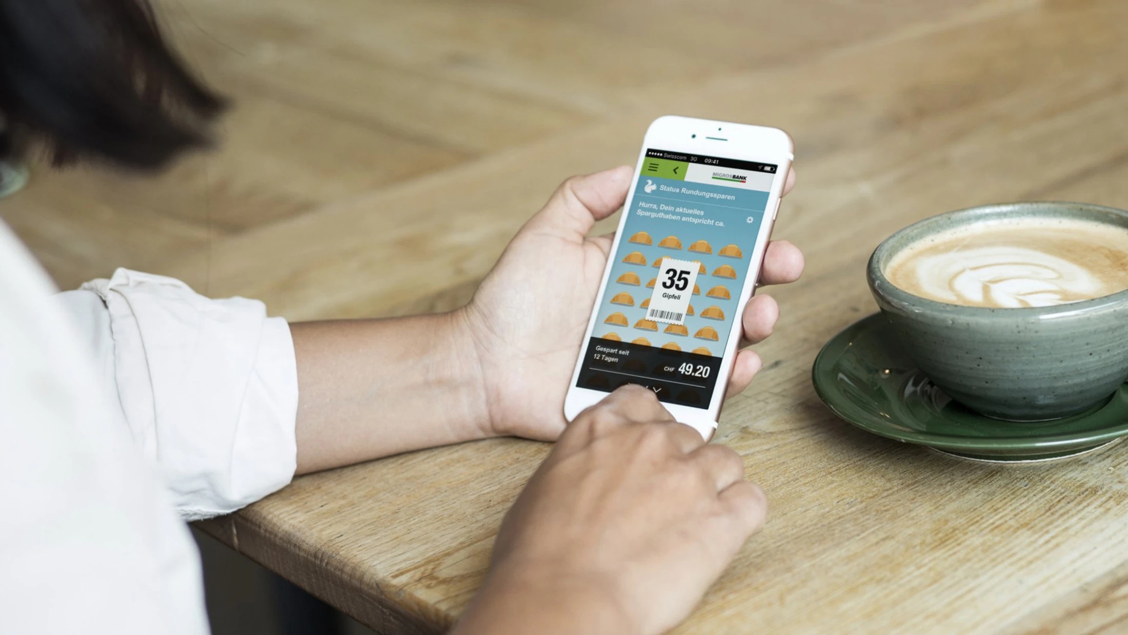 Das Bild zeigt eine Person, die an einem Holztisch sitzt und ein Smartphone in der rechten Hand hält. Auf dem Smartphone ist die mobile Banking-App der Zuger Kantonalbank geöffnet. Der Bildschirm zeigt eine Sparfunktion, die visuell ansprechend dargestell
