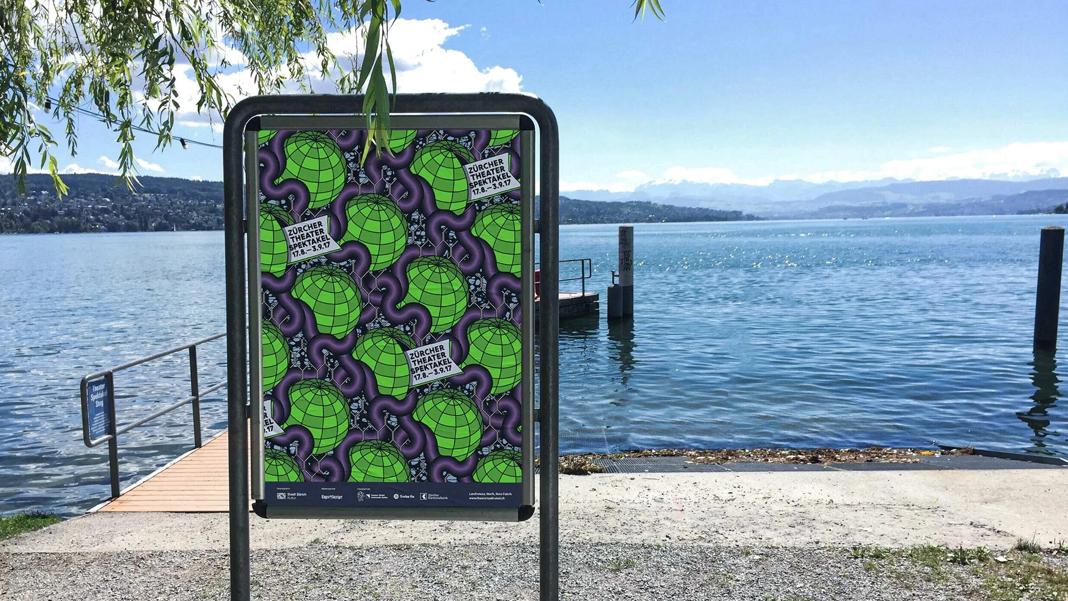 Das Bild zeigt ein Plakat für das Zürcher Theaterspektakel, das an einem metallischen Rahmen nahe einem See aufgestellt ist. Im Hintergrund ist ein See mit klarem blauem Wasser zu sehen, der von einer malerischen Landschaft und entfernten Bergen umgeben